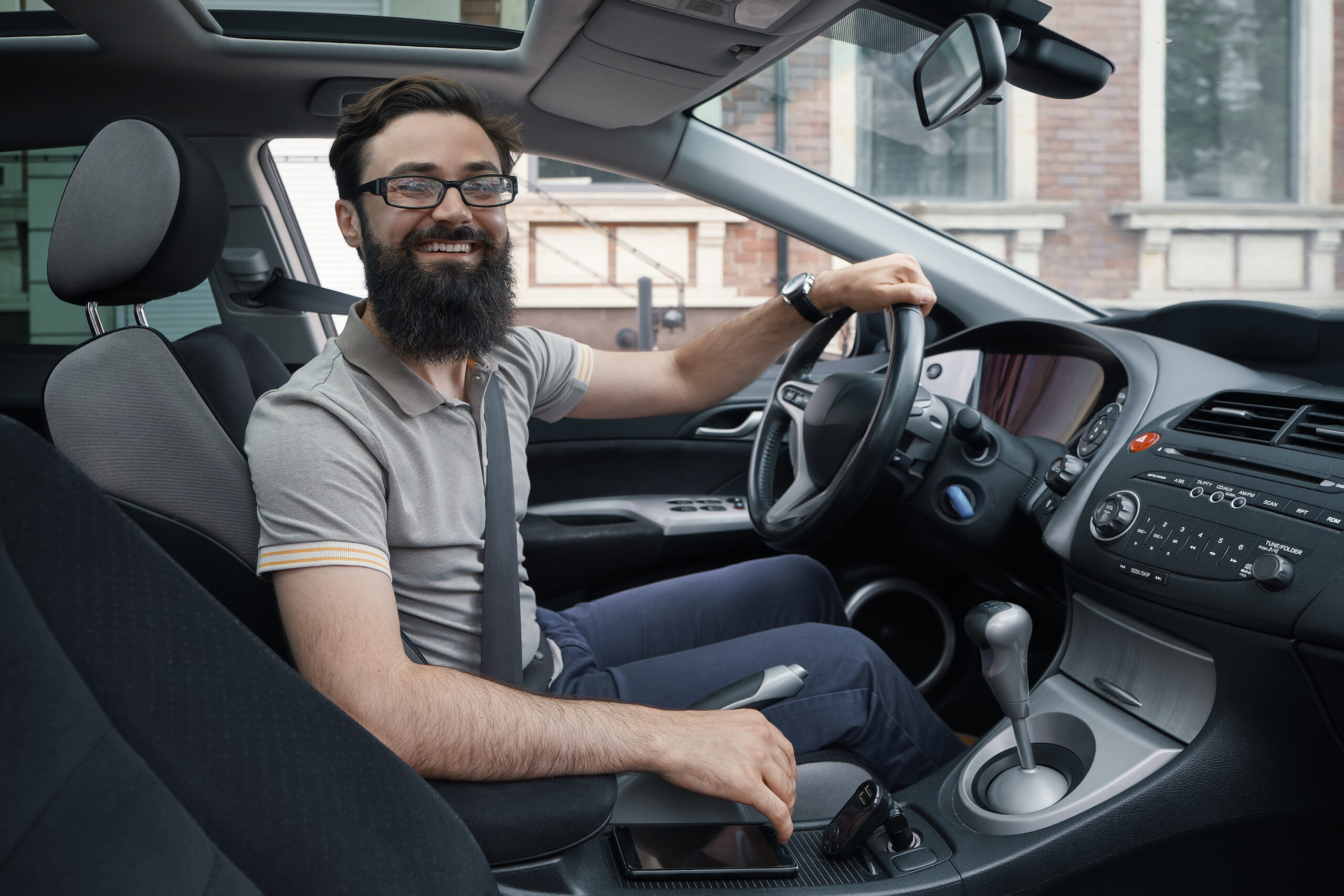 I love my car. A positive bearded man driving his car through the city while smiling. Enjoying every day simple moments. Side view